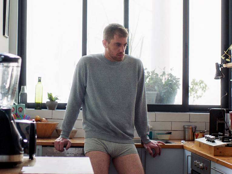 Eine männlich gelesene Person steht in grauem Pullover und Unterhose an eine Theke gelehnt in der Küche. Hinter ihm ist ein Fenster, die Person scheint in Gedanken versunken.